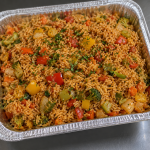 Half-size aluminum catering tray filled with vegetarian Cajun jambalaya featuring seasoned rice, bell peppers, onions, broccoli, carrots.