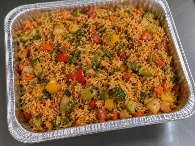 Half-size aluminum catering tray filled with vegetarian Cajun jambalaya featuring seasoned rice, bell peppers, onions, broccoli, carrots.